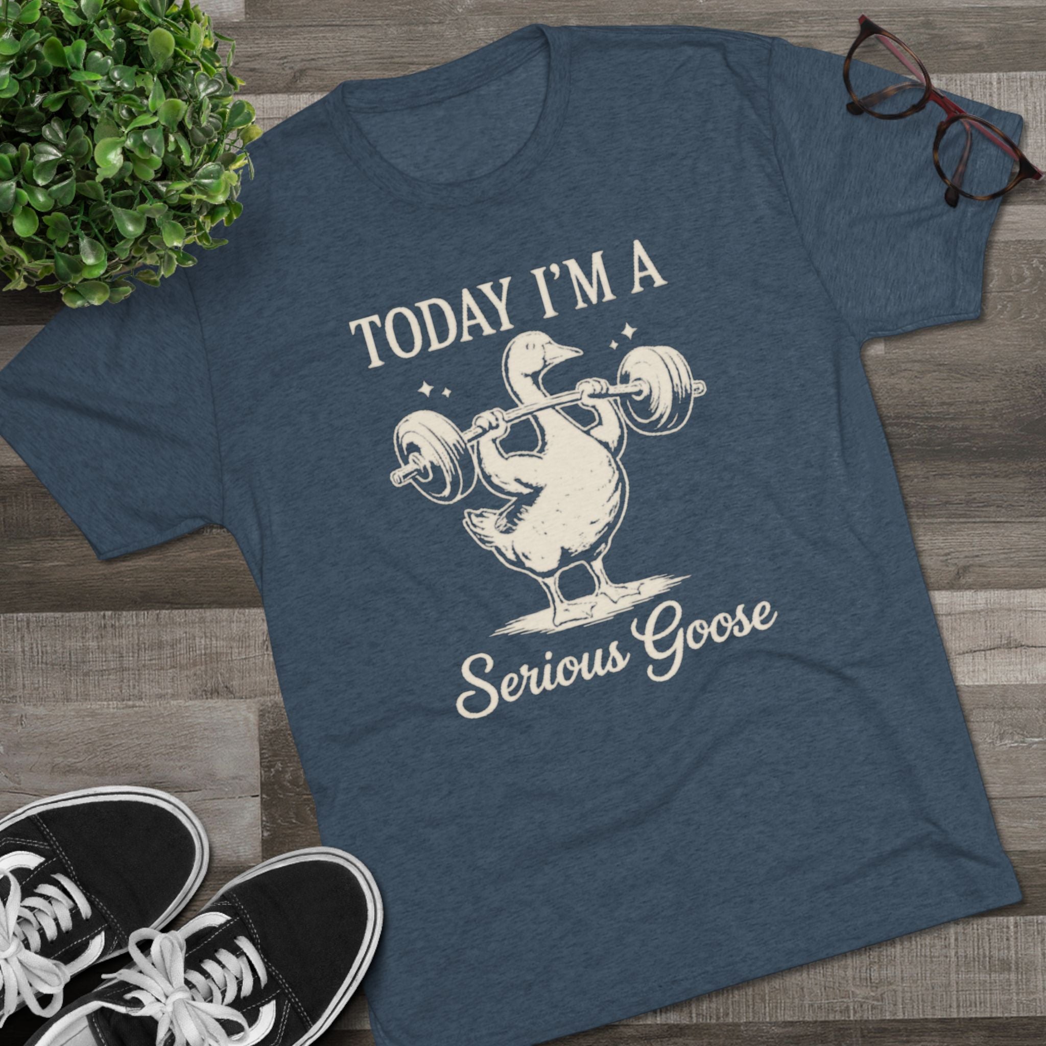 Blue t-shirt with a graphic of a goose lifting weights and text 'Today I'm a Serious Goose' on a wooden surface with sneakers and a plant.