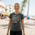 Woman wearing a t-shirt with 'Lift Heavy Pray Hard' text on a beachside boardwalk.
